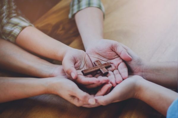 Three people hands holding small wooden cross on wood table, Christian concept Jesus is the center of life or ministry , copy space.